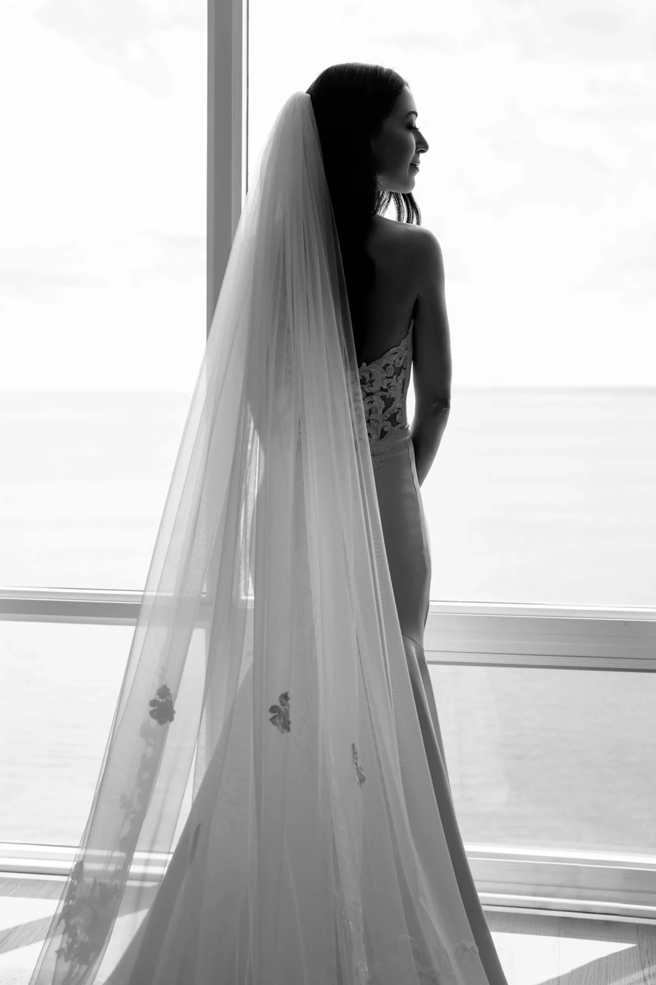 Black and white bridal portrait showing a bride standing by a window with her veil flowing behind her