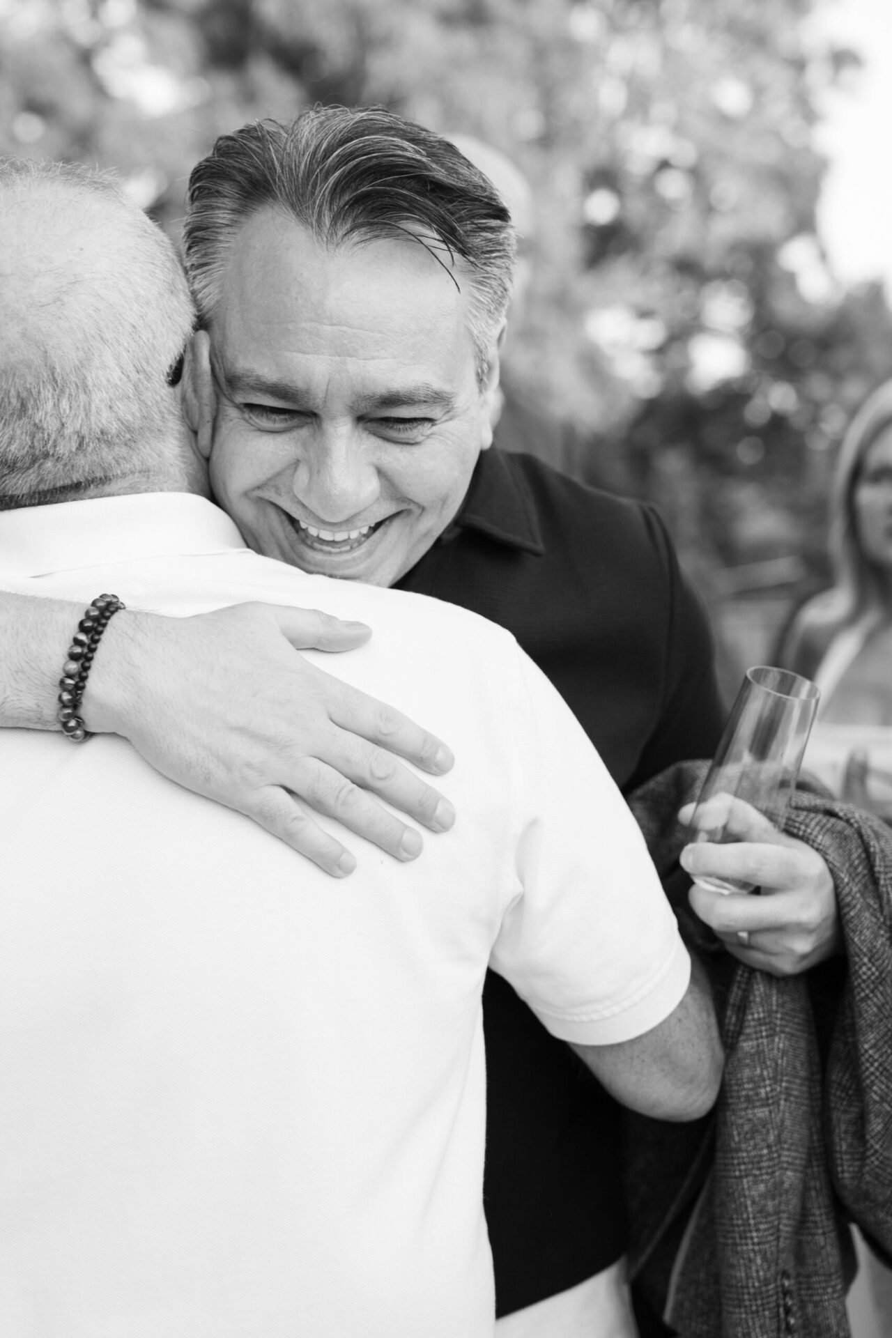 Black and white candid photograph of a smiling man hugging another guest during a celebration