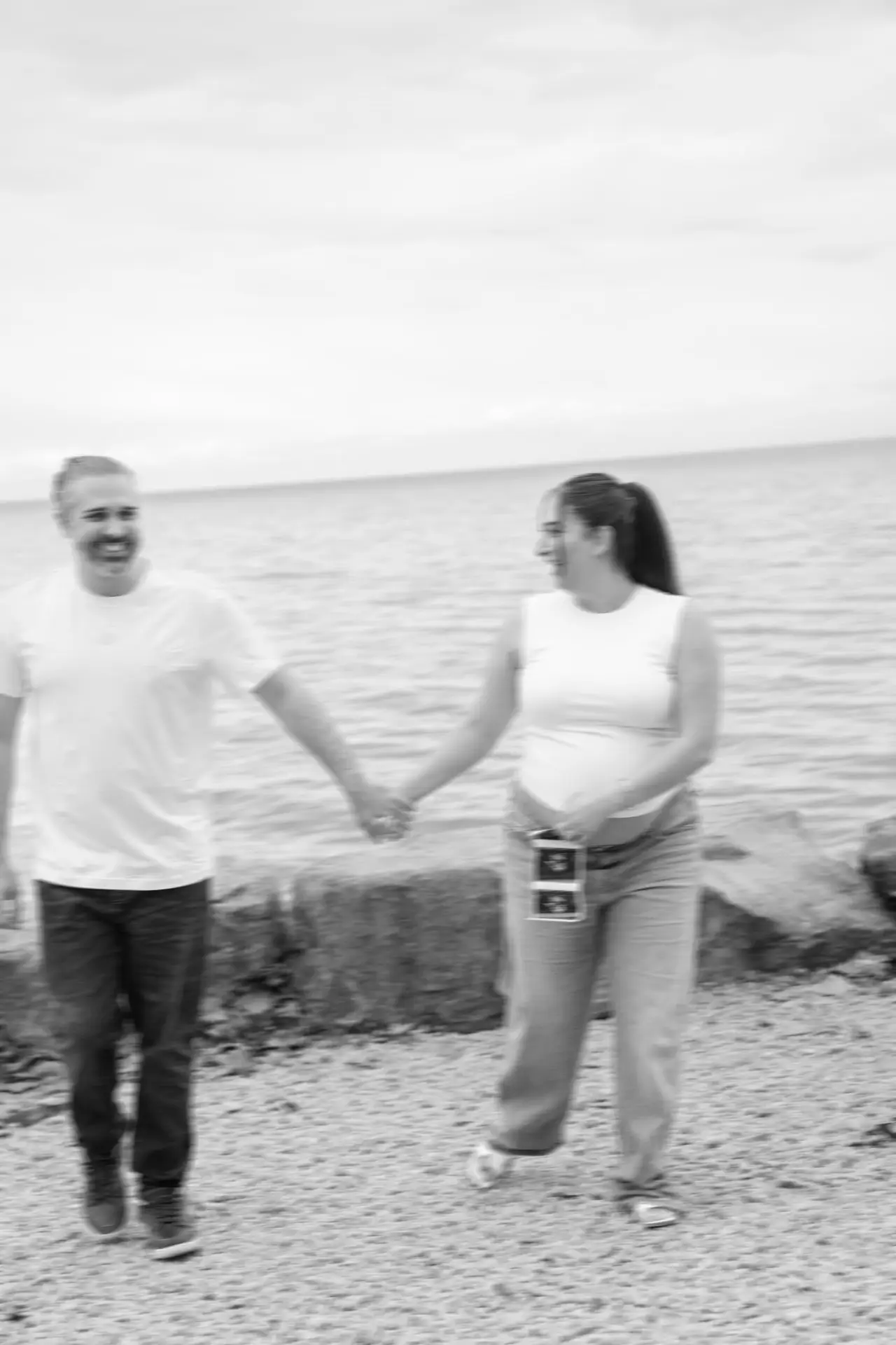 Black and white photograph of an expecting couple holding hands together by the water